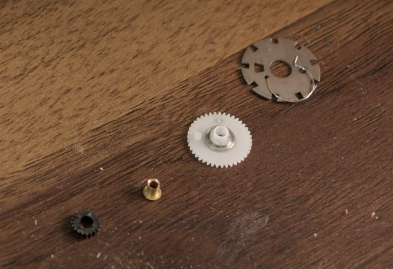 Disassembled stock frame spacing assembly parts laid out on a table: black gear, brass bushing, white gear, and OEM frame spacing disc.